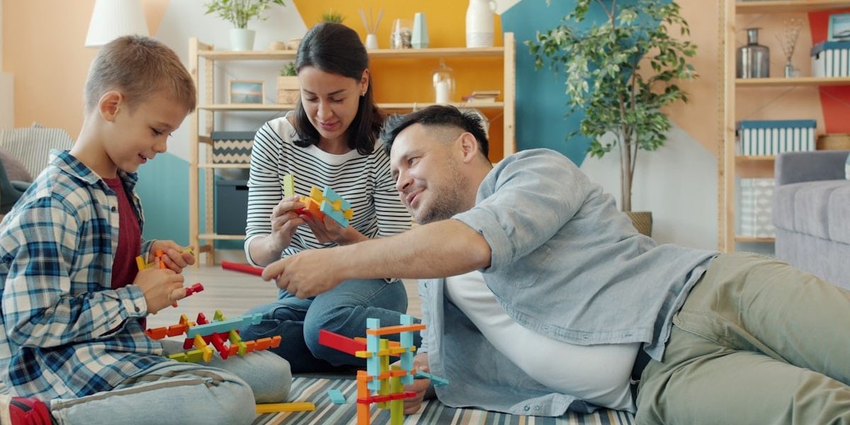 Child is playing with mother and father cheerful adults at home building with construction blocks sitting on floor together. Youth and family concept.