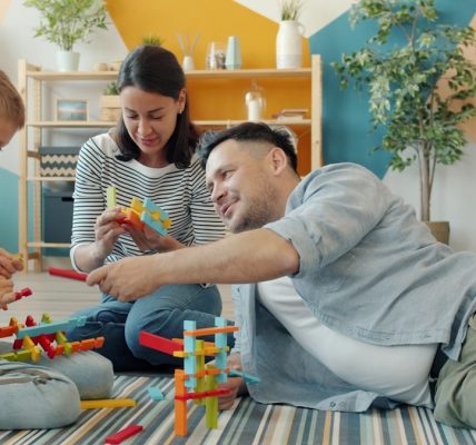 Child is playing with mother and father cheerful adults at home building with construction blocks sitting on floor together. Youth and family concept.