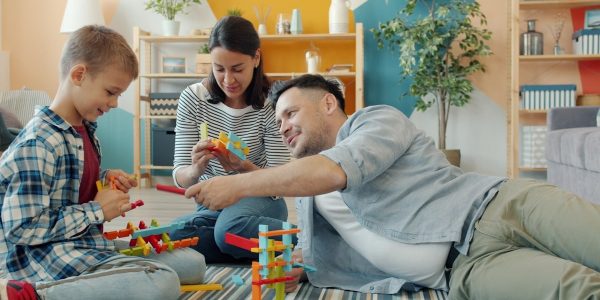 Child is playing with mother and father cheerful adults at home building with construction blocks sitting on floor together. Youth and family concept.